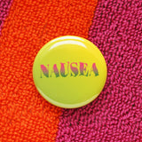 Round pinback button with yellow to green ombre background, with pink to green ombre text that reads NAUSEA in a wavy serif font. Button is on a bright orange and magenta Terry cloth robe.