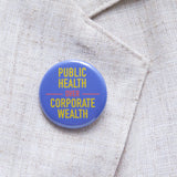 A round pinback button with a scrubs blue-purple background that reads PUBLIC HEALTH OVER CORPORATE WEALTH in yellow and neon red. Button is on a cream blazer lapel.