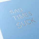 Powder blue greeting card that says SAD TIMES SUCK. Close up of the silver hot foil pressed text.