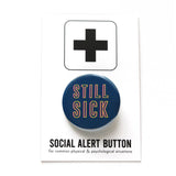 Dark blue round pinback button with neon yellow & pink text that reads STILL SICK. Button is on a Social Alert Button backing card.