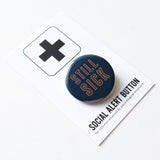 Angled, dark blue round pinback button with neon yellow & pink text that reads STILL SICK. Button is on a Social Alert Button backing card.