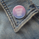 Light pink & blue, round, pinback button that reads SUPPORT TRANS KIDS. Button is pinned to the collar of an old denim jean jacket.