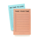 Two vertical rectangle notepads stacked  with a peach background notepad on the top, reading TAKE YOUR TIME in black text at the top, with left justified check boxes and lines to write on to the right. The brand's website is listed small at the bottom of the notepad: www.wordforwordfactory.pro. A light blue matching notepad is beneath looking identical except for the header NOT TIME TO LOSE on a light blue background.