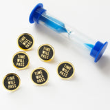 Five round TIME WILL PASS enamel pins next to a small hour glass.