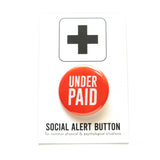 Bright round red-orange pinback button that reads UNDER PAID in white san serif text. Pinned to a Social Alert Button backing card.