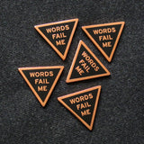 Five WORDS FAIL ME enamel pins on a gray felt background.