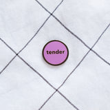 Round enamel pin that says TENDER on fabric background.