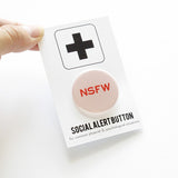 A round light pink pinback button with dark pink text that reads NSFW stands for Not Safe For Work. Button is on a Social alert button backing card, held by a hand.